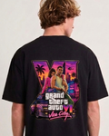 A man wearing a black t-shirt, viewed from the back. The shirt features a large graphic print of the video game Grand Theft Auto VI, showing characters and the game's logo in vibrant neon colors over a sunset cityscape.