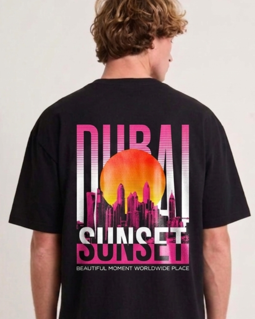 A man viewed from behind wearing a black t-shirt with a large graphic print on the back featuring a sunset over a city skyline in pink, orange, and white, with the text 'DUBAI SUNSET'.